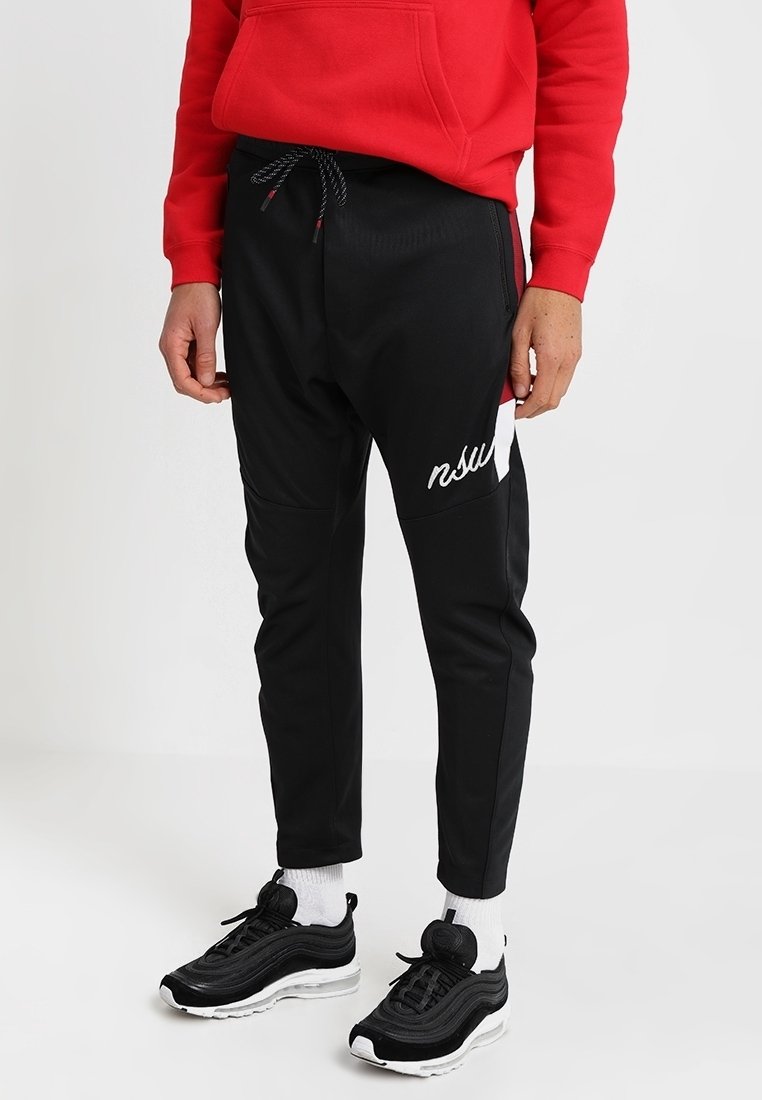 Persoon in zwarte joggers metkoord en witte tekst op de dij, witte sokken, zwart-witte sneakers en een rode hoodie.