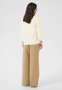 Grietinė bluzonė su gėlių tekstūra ir pūstomis rankovėmis, derinama su aukštu juosmeniu, plačiomis bežinėmis kelnėmis, rodoma iš nugaros.