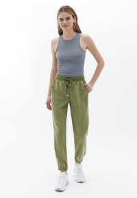 Graues, eng anliegendes Tanktop und olivgrüne Jogginghosen mit elastischen Bündchen und Kordelzug. Modell trägt weiße Sneakers. Minimalistisches Design.