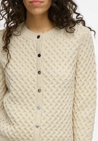 Cárdigan de punto beige con un patrón texturizado, que presenta un cuello redondo y botones negros en la parte delantera.