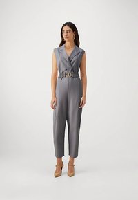 Kvinne i grå ermeløs jumpsuit med belte og beige hæler, står mot en lys bakgrunn og ser til siden.