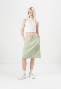 Világos zöld vászon szoknya, amely pamutból készült, két első zsebe van, gombos zárása és egyenes fazona. Fehér, cropped tank top-mal viselve.