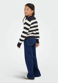 Chica sonriendo sobre el hombro, vistiendo un suéter a rayas azul marino y blanco, jeans de pierna ancha y zapatillas blancas, de pie contra un fondo sencillo.