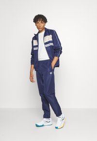 Navy tracksuit made of smooth material with light beige accents and white stripes. Paired with white sneakers featuring turquoise and yellow details.