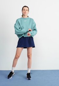Sudadera verde azulado, falda de rayas navy, calcetines blancos y zapatillas negras. El conjunto tiene un corte relajado y un logo sutil en la sudadera.