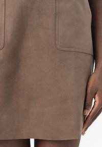 Jupe en daim marron avec deux poches avant, texture lisse et design épuré, tombant au-dessus du genou, maintenue par une main sur le côté.