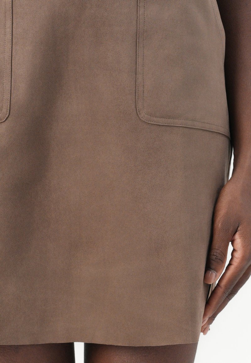 Falda de ante marrón con dos bolsillos frontales, textura suave y un diseño limpio, que cae por encima de la rodilla, sostenida por una mano a un lado.