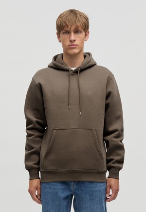 Joven con cabello rubio que lleva una sudadera marrón con capucha y vaqueros azules, de pie frente a un fondo claro y liso.