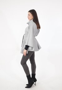 Grijze, gestructureerde cape-stijl jas met een hoge kraag, een ceintuurlijn en een gelaagde zoom. Draagt over een zwart shirt, gecombineerd met grijze jeans en zwarte laarzen.