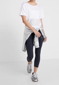Vrouw die loopt in een witte shirt met korte mouwen, een zilveren jack om haar taille, zwarte cropped leggings en grijze hardloopschoenen.