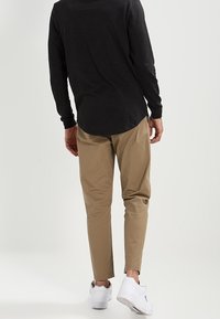 Mężczyzna stojący tyłem, ubrany w czarną bluzę z długim rękawem, beżowe spodnie i białe trampki na jednolitym tle.