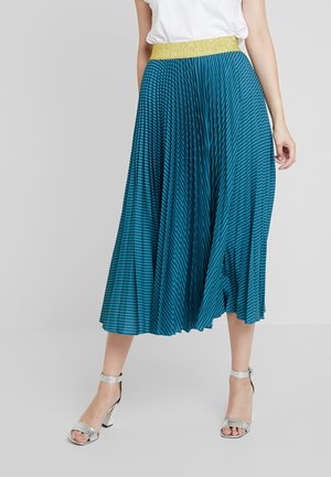 Femme portant une jupe midi plissée à rayures bleu sarcelle et noires avec une ceinture scintillante dorée et des sandales à talons hauts argentées.