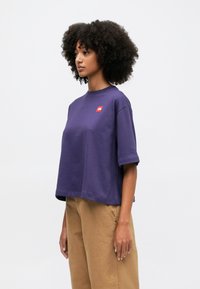 Donna con capelli ricci che indossa una maglietta corta viola larga e pantaloni beige, in piedi di lato su uno sfondo chiaro e semplice.