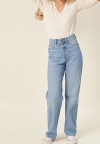 Femme portant un jean taille haute bleu clair, des baskets blanches et un haut à manches longues crème, soulevant une veste beige sur ses épaules.