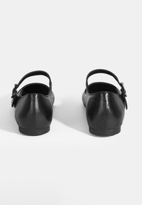 Schwarze Lederschuhe mit runder Zehenform und einem Schnallen-Riemen am Knöchel. Glatte Textur und flache Sohle. Die Rückansicht zeigt ein minimalistisches Design.