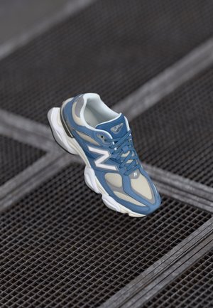 Blue and beige New Balance sneaker with white sole, floating above a metal grid surface.