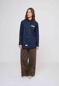 Nő hosszú tengerészkék gombos ingben, díszített zsebbel és túlméretezett barna kordbársony nadrágban, egyszínű világos háttér előtt állva.