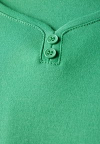 Zelený krátký rukáv top s henley výstřihem a dvěma odpovídajícími knoflíky, vyrobený z měkké texturované látky. Viditelné jemné švy.
