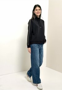 Sweatshirt noir à col montant avec des rayures blanches sur les côtés, associé à un jean bleu ample et des baskets blanches. Tissu lisse et coupe décontractée.
