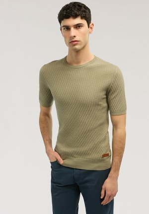 Junger Mann, der einen strukturierten, olivgrünen Pullover mit kurzen Ärmeln und dunkelblaue Hose trägt, mit einer Hand in der Tasche vor einem einfarbigen Hintergrund steht.