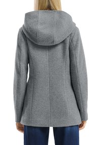 Femme portant une veste à capuche texturée grise avec des manches longues, vue de dos, associée à un jean bleu foncé.