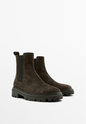 Bruine suede enkelbotten met een stevige zwarte rubberen zool, elastische zijkanten en lusjes voor gemakkelijk aan- en uittrekken.