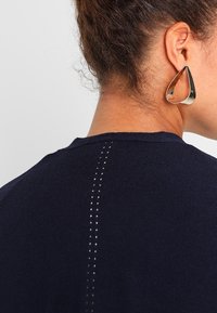 Marinblå tröja med ribbad textur, prydd med en vertikal rad av små, ljusa dekorationhål längs ryggen. Silverfärgade droppformade örhängen.