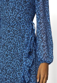 Modrá květinová šaty vyrobené z lehké látky, s řaseným lemem, dlouhými rukávy a mašlí v pase. Černý květinový vzor po celém povrchu.