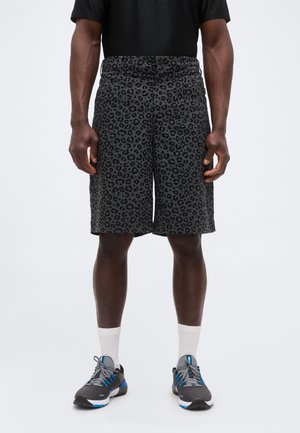 Mężczyzna noszący czarne i szare spodenki w cętki lamparta, czarną koszulkę, białe skarpetki i szare buty sportowe z niebieskimi akcentami, stojący na białym tle.