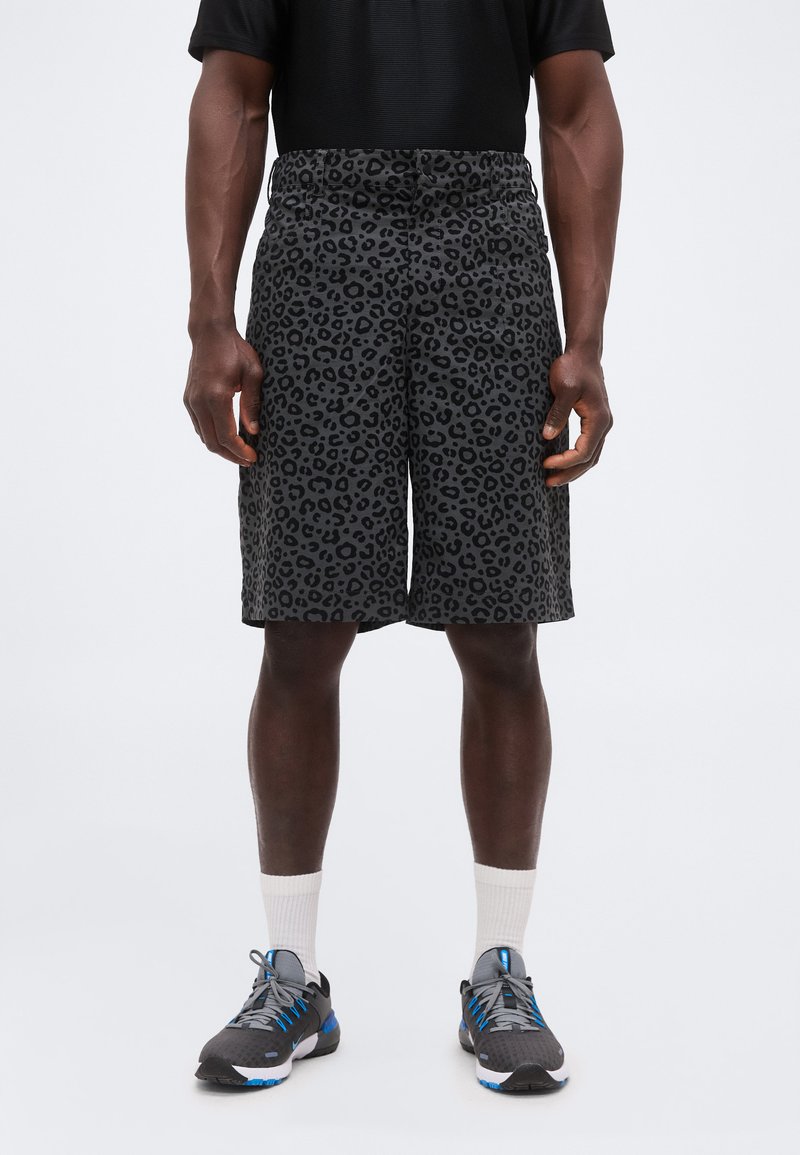 Άνδρας που φορά μαύρα και γκρι σορτς με σχέδιο λεοπάρδαλης, μαύρο πουκάμισο, λευκές κάλτσες και γκρι αθλητικά παπούτσια με μπλε λεπτομέρειες, στέκεται μπροστά σε λευκό φόντο.