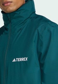 Veste imperméable teal avec col montant et fermeture éclair devant. Présente le logo blanc "TERREX" sur la poitrine gauche. Texture lisse et légère.