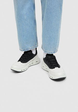 Osoba nosząca jasnoniebieskie dżinsy, białe skarpetki oraz czarno-białe masywne sneakersy stojąca na białej podłodze.