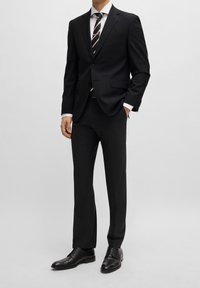 Completo nero con giacca aderente e pantaloni dritti, abbinato a una camicia bianca e una cravatta a righe. Le scarpe eleganti in pelle nera completano il look.