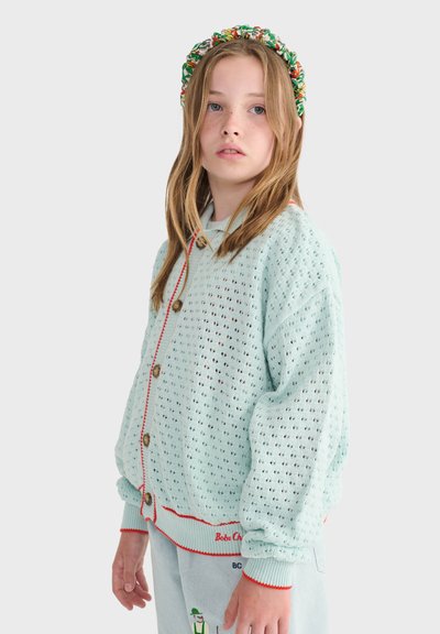 Fille aux longs cheveux portant un cardigan bleu clair à mailles ouvertes avec boutons et bordure rouge, pantalon clair et serre-tête fleuri coloré.