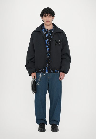 Jachetă puffer neagră, cămașă florală, pantaloni din denim albastri cu embellishments, pantofi negri și o geantă mică neagră. Prezintă un detaliu unic în zona buzunarului de la piept.