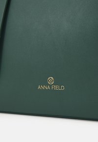 Grøn taske i imiteret læder med en glat tekstur, der har en guldfarvet "ANNA FIELD"-logo og et minimalistisk design.