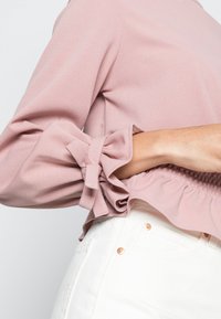 Donna che indossa una blusa rosa a maniche lunghe con polsini annodati e pantaloni bianchi a vita alta con dettagli in rivetti di rame.