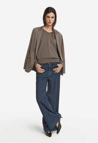 Brauner Strickpullover unter einem beigen Blazer, kombiniert mit weit geschnittenen, dunklen Jeans. Schwarze Absätze runden das Outfit ab.