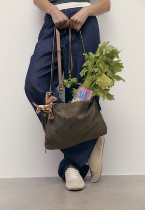 Persona che indossa jeans e sneakers beige, mentre tiene una borsa marrone contenente sedano, una rivista e un portachiavi a forma di cane, su uno sfondo neutro.