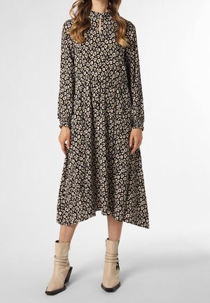 Frau trägt ein langärmliges, knielanges Kleid mit Leopardenmuster, kombiniert mit beigen Stiefeletten, vor einem schlichten Hintergrund.