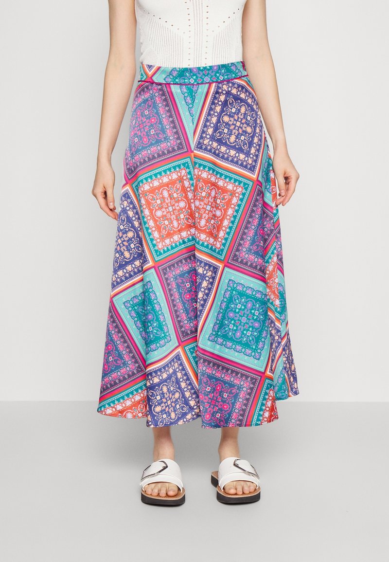 Žena oblečená v barevné midi sukni s geometrickými a květinovými vzory, doplněná bílými sandály a bílým topem bez rukávů.