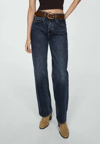 Jeans de pierna recta azul oscuro con un cinturón marrón, que cuentan con cinco bolsillos y detalles de costura sutiles. Modelo combinado con botas de tacón color beige.