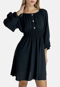 Schwarzes Kleid aus leichtem Stoff, mit gesmokter Taille, langen Puffärmeln und drei großen Knöpfen an der Vorderseite.