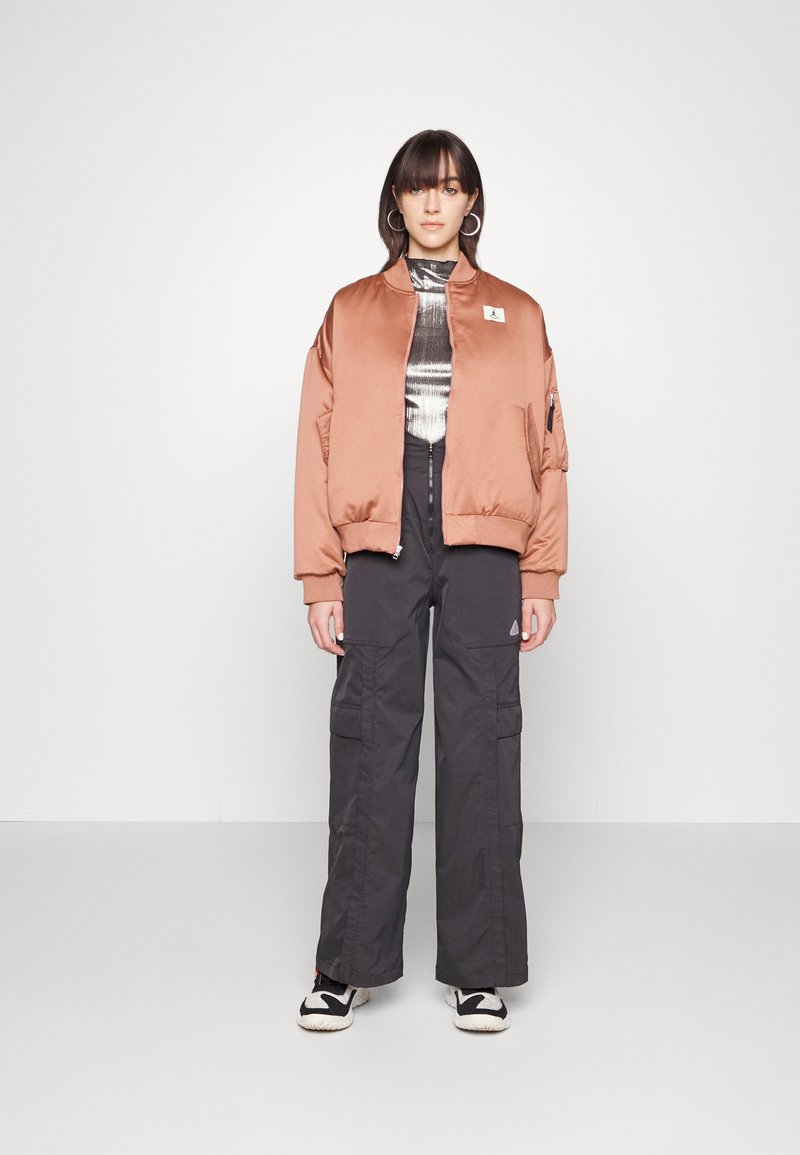 Veste bomber en bronze avec une texture lisse, fermeture éclair frontale et poches latérales, associée à un pantalon cargo foncé et un haut à motifs, complétée par des baskets noires.