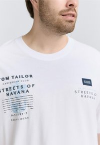 Man in een wit T-shirt met de tekst "Streets of Havana" en "Tom Tailor Caribbean Club" en een blauwe grafische afbeelding op de voorkant.