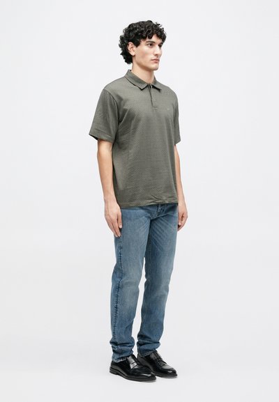 Polo vert à motifs avec col, associé à un jean bleu. Le mannequin est debout, portant des chaussures habillées noires. Design et coupe simples.