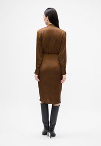 Abito midi in satin marrone con motivo floreale, maniche lunghe e vita cinte. Il modello indossa stivali al ginocchio neri per creare un contrasto.