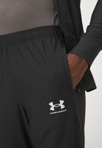 Čierne športové nohavice s elastickým pásom, so bočnou vreckom a bielym logom Under Armour na dolnej časti nohy. Hladká, ľahká tkanina.