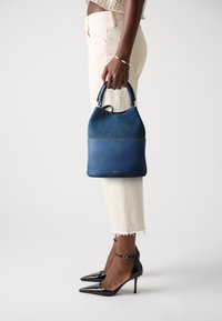 Blue leather and suede handbag with a rounded top, featuring a short handle and contrasting stitching. Paired with light-colored pants and black heels.