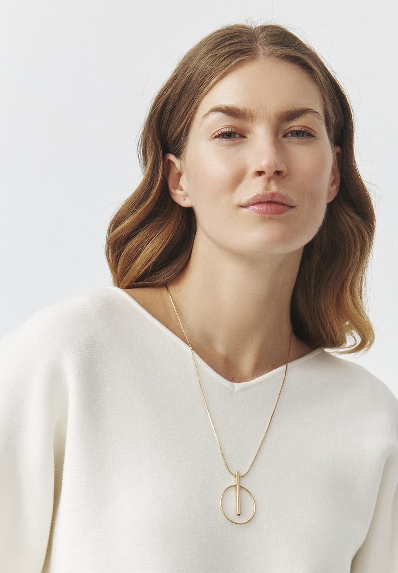Gul vedhængs halskæde med en lodret stang og cirkulær accent, båret af en model iført en hvid V-hals sweater. Glat tekstur.